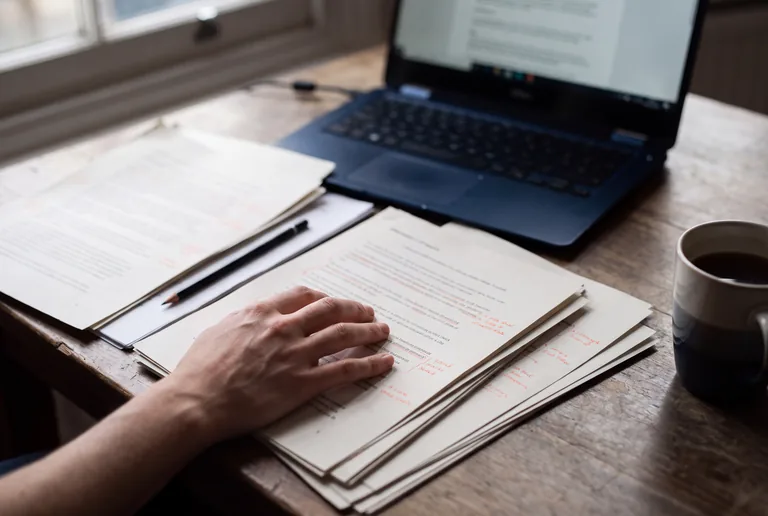 Printed article drafts marked up by hand on a desk beside a laptop
